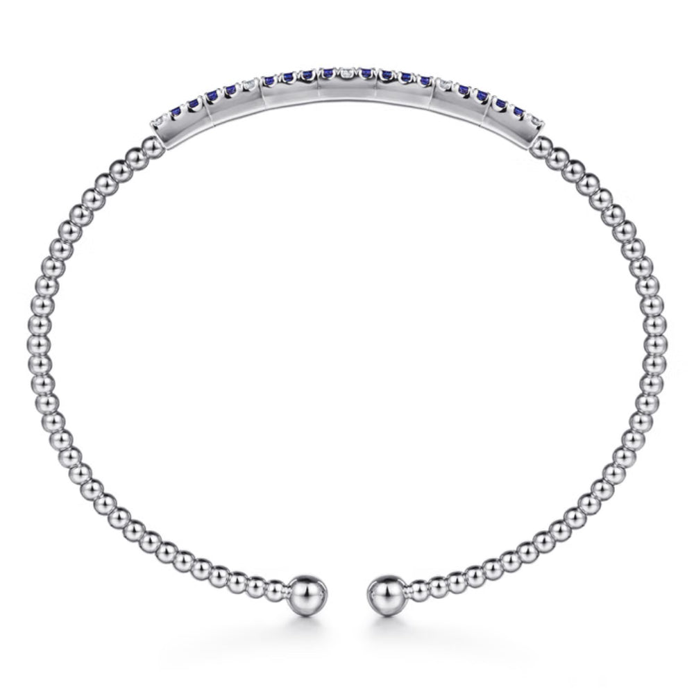 A silver-toned beaded open cuff bracelet featuring a curved top band set with small dark blue gemstones, displayed on a clean white background.