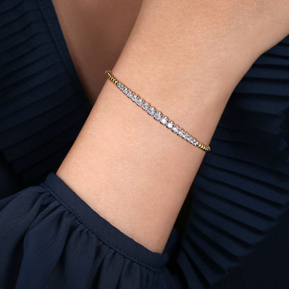 A delicate gold tennis bracelet with clear cubic zirconia stones worn on a person's wrist, set against a dark navy blue pleated top.