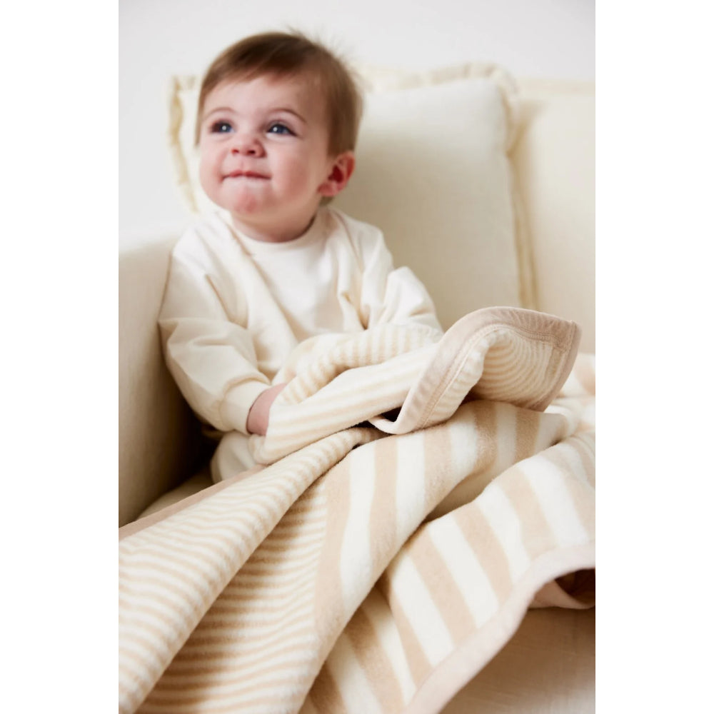 A baby wearing a cream long-sleeve bodysuit sits on a beige sofa, wrapped in a cream and beige striped blanket.
