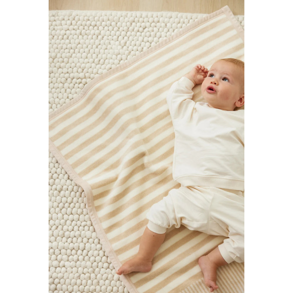 A baby in a cream-colored onesie and pants lies on a cream and beige striped blanket, which is laid over a textured cream rug.
