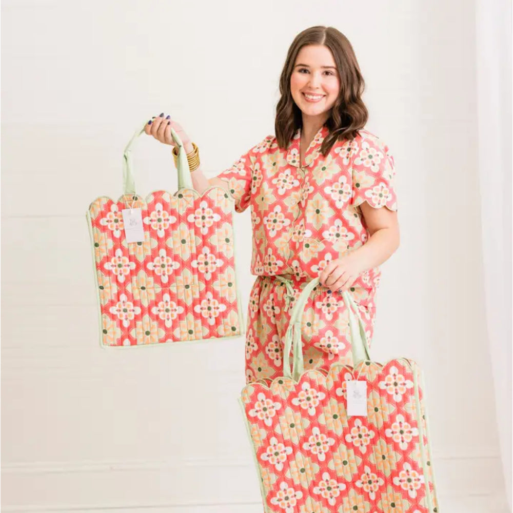 A woman wearing a matching coral and beige patterned short-sleeve shirt and shorts holds two large fabric tote bags with the same geometric print and light green handles against a white paneled wall.
