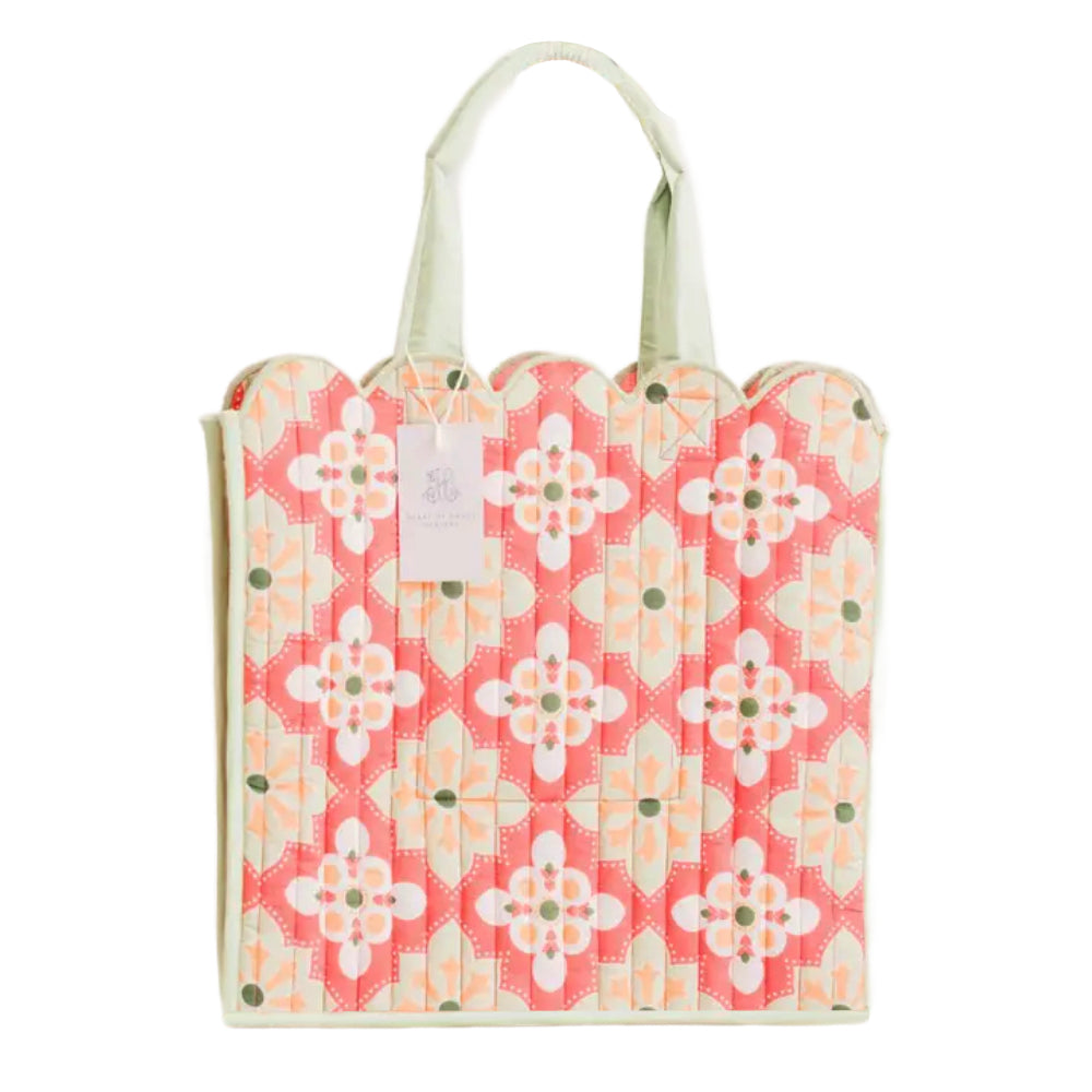 A quilted fabric tote bag with a coral pink and cream floral ikat pattern, featuring light green handles and a hanging paper tag.
