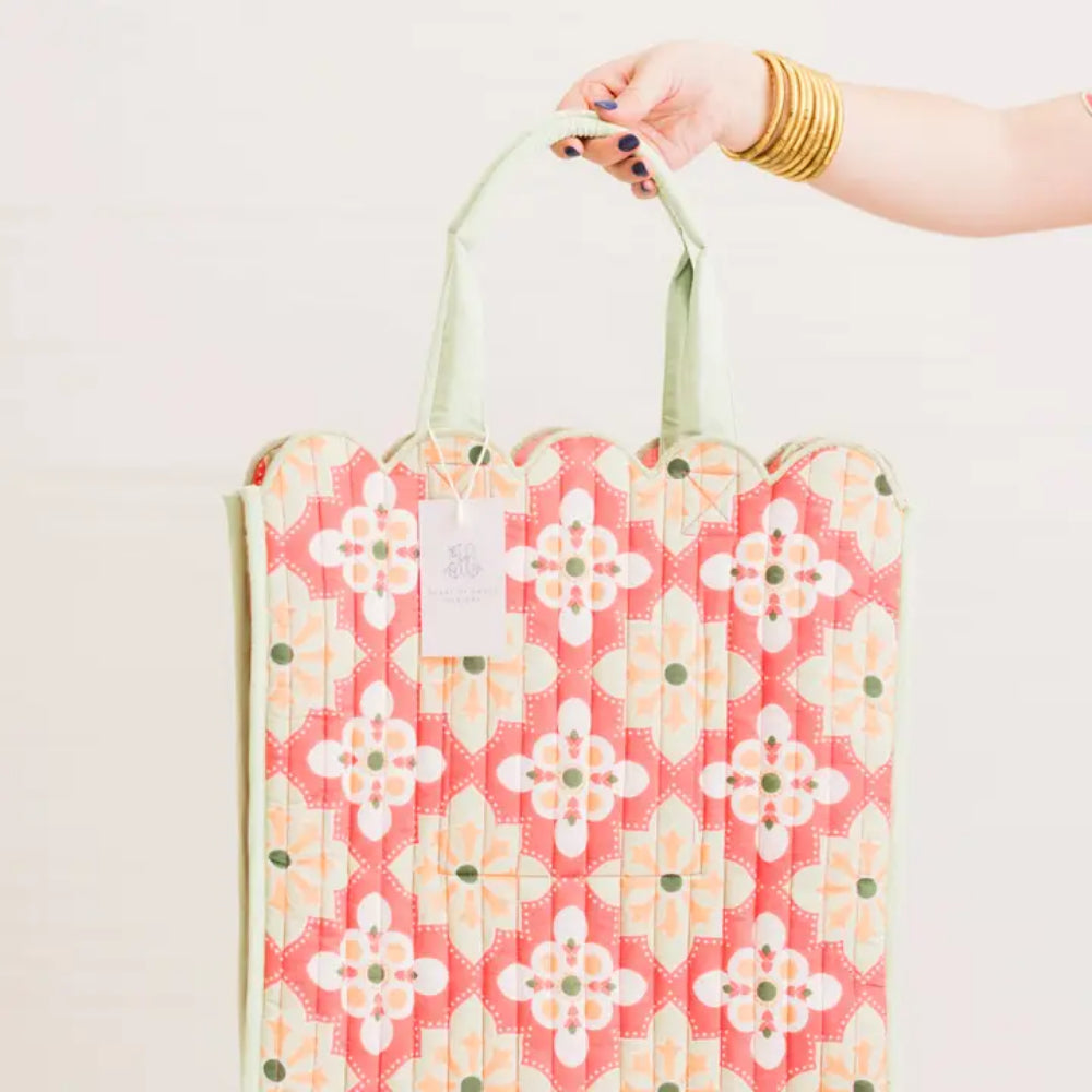 A quilted shopping tote with a coral pink, white, and yellow floral pattern and light green handles, held by a person's hand against a plain white background.