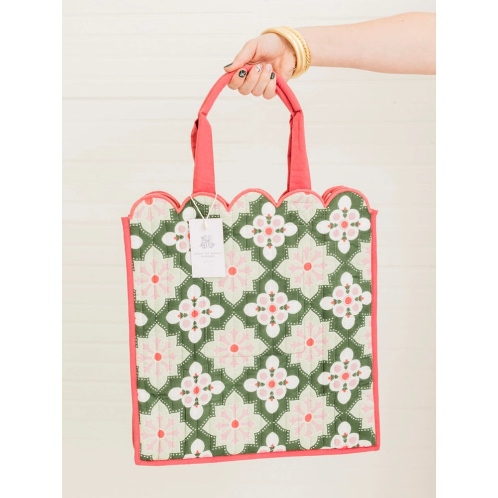 A hand holding a green and pink patterned fabric tote bag with scalloped edges and coral handles, set against a light wood paneled background.