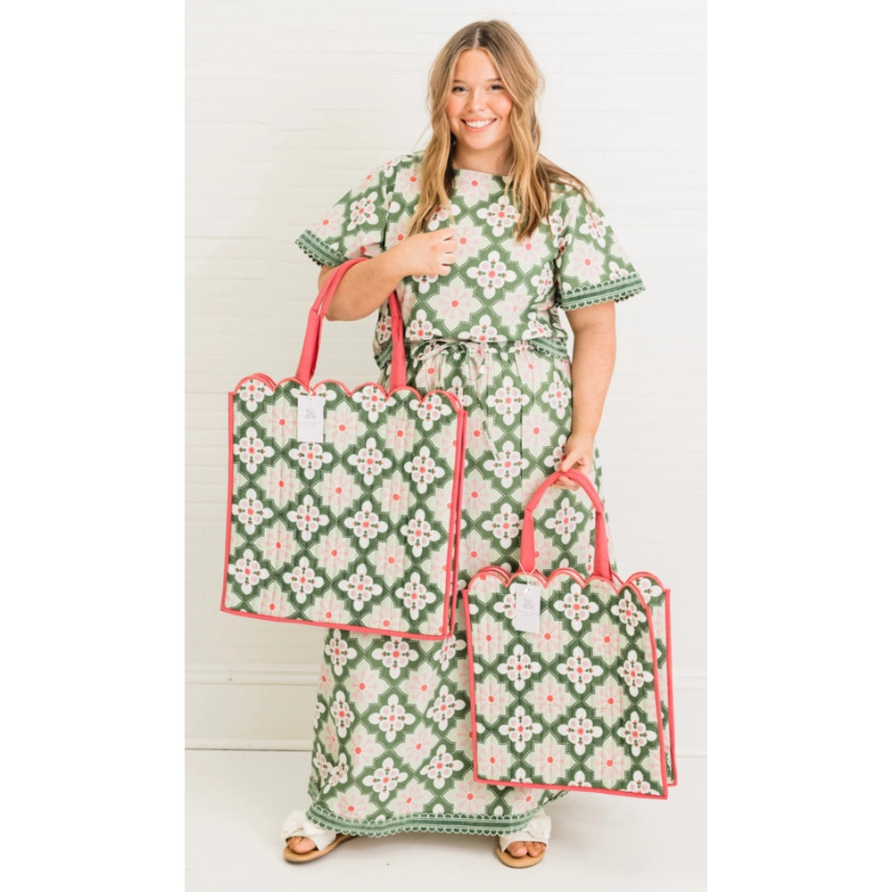 A woman stands against a white paneled wall holding two matching tote bags. She is wearing a matching green and pink patterned dress.