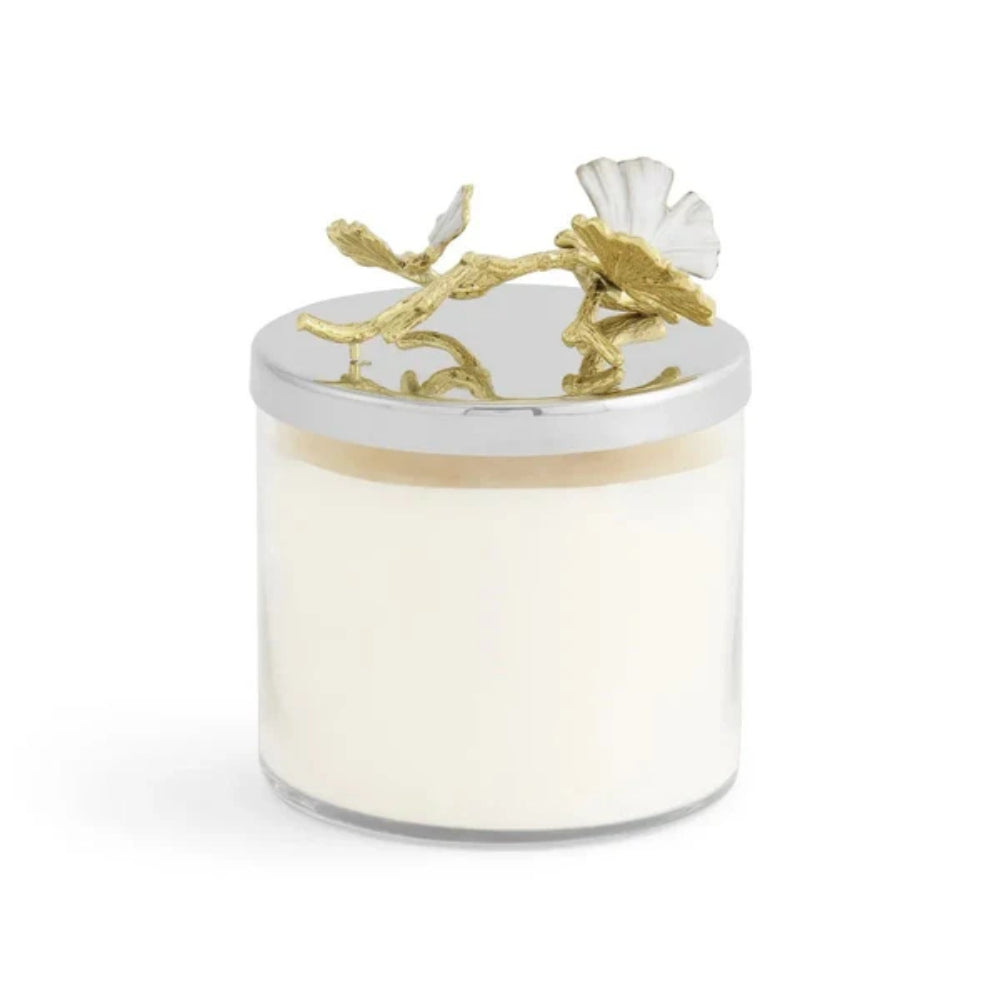 A clear glass jar candle filled with creamy off-white wax, featuring a polished silver metal lid adorned with a sculpted gold branch and white flower motif, set against a plain white background.