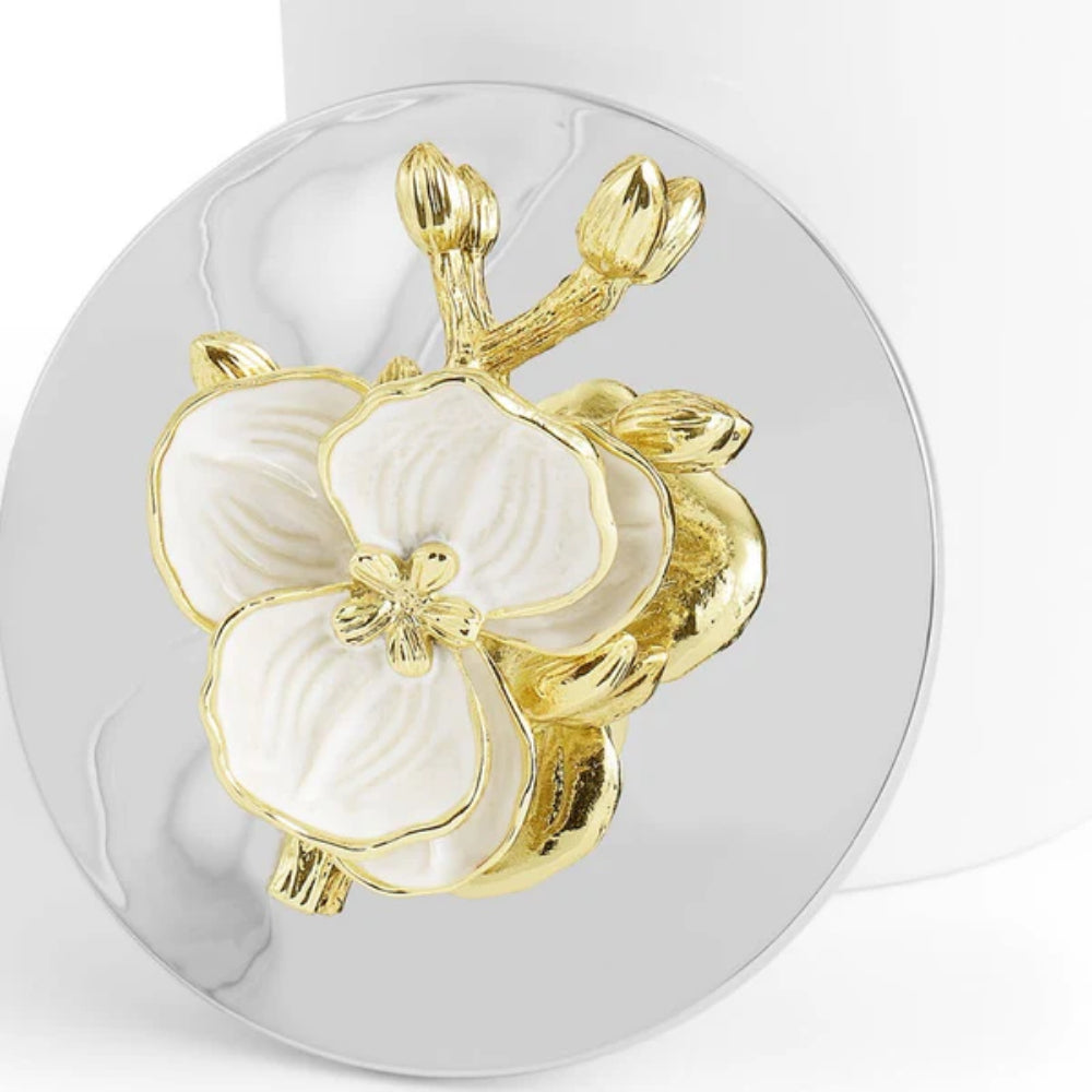 A close up photo of the lid of a small white candle with a white and gold floral design on a silverplated lid on a white backround