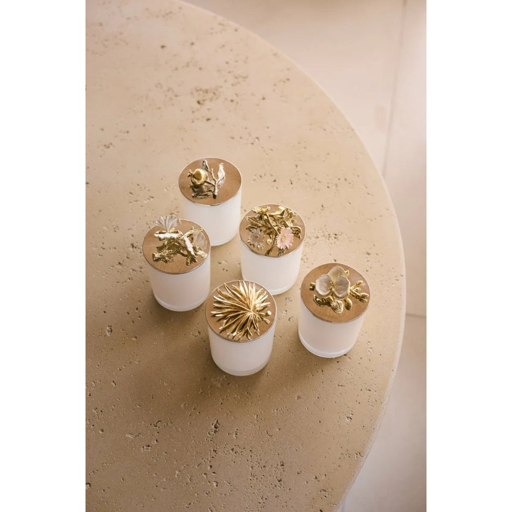 Five small white cylindrical containers with metallic gold lids arranged on a round beige stone table. Each lid features a different decorative gold floral or starburst design.