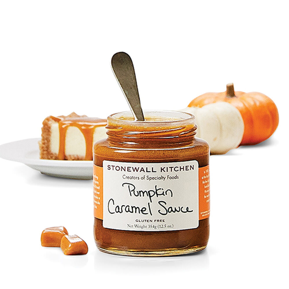 Jar of Stonewall Kitchen Pumpkin Caramel Sauce with a spoon, surrounded by pumpkins and a slice of cake on a white background.