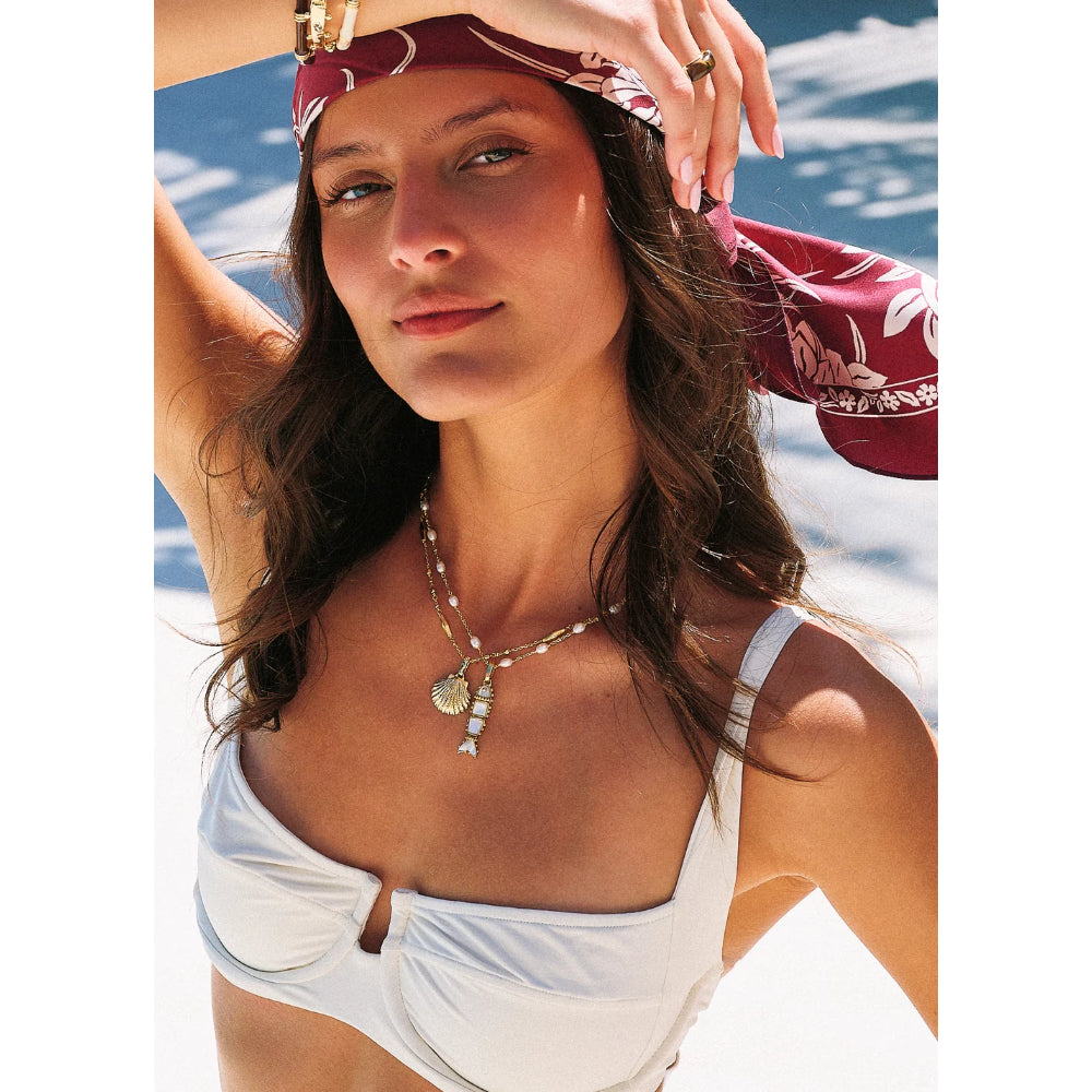 A woman outdoors wearing a white bikini top and a red patterned headscarf, with a bohemian-style silver pendant necklace featuring a seashell charm.