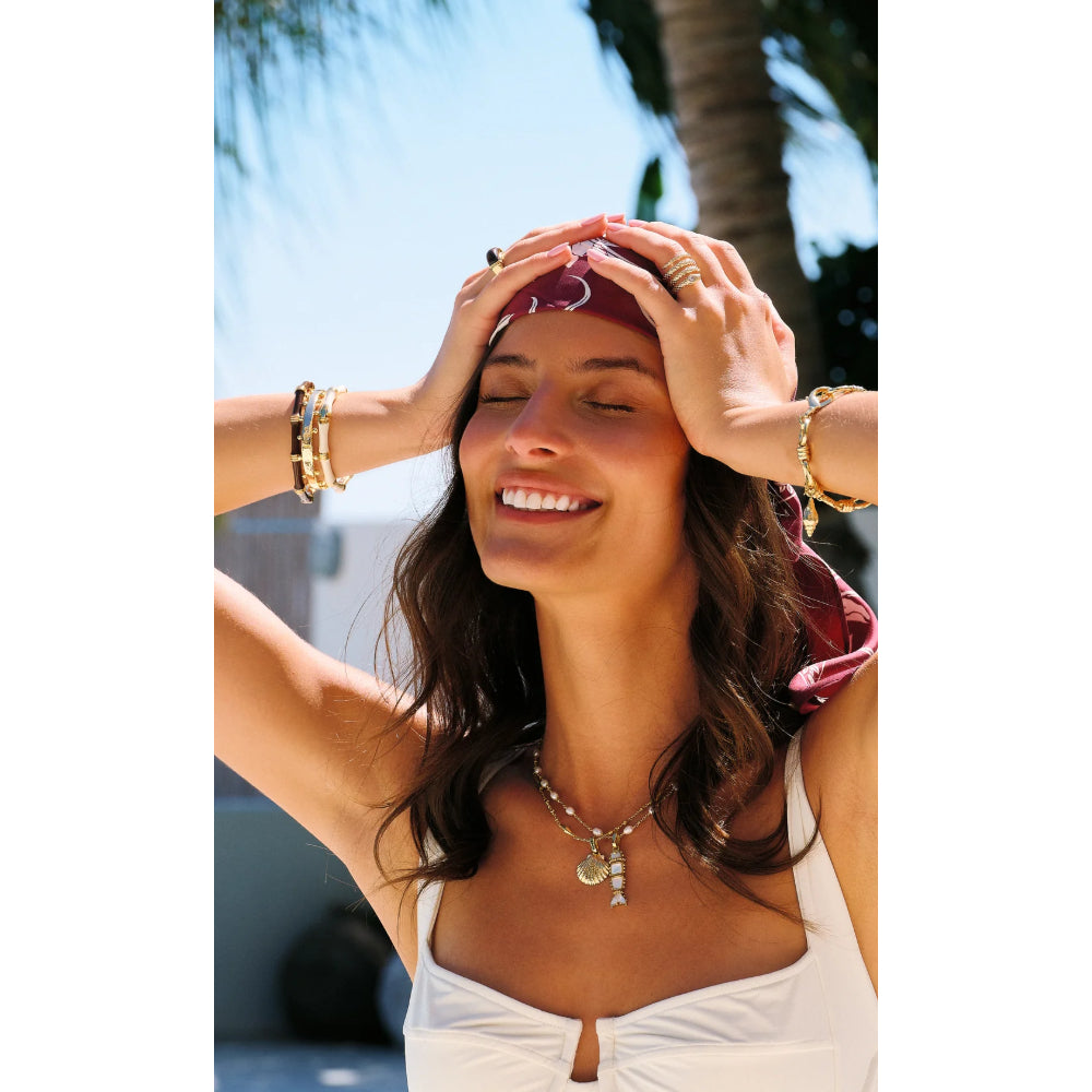 A woman outdoors wearing a white bikini top and layered gold necklaces and bracelets, shielding her eyes from the sun with her hands. Palm trees and blue sky are visible in the background.