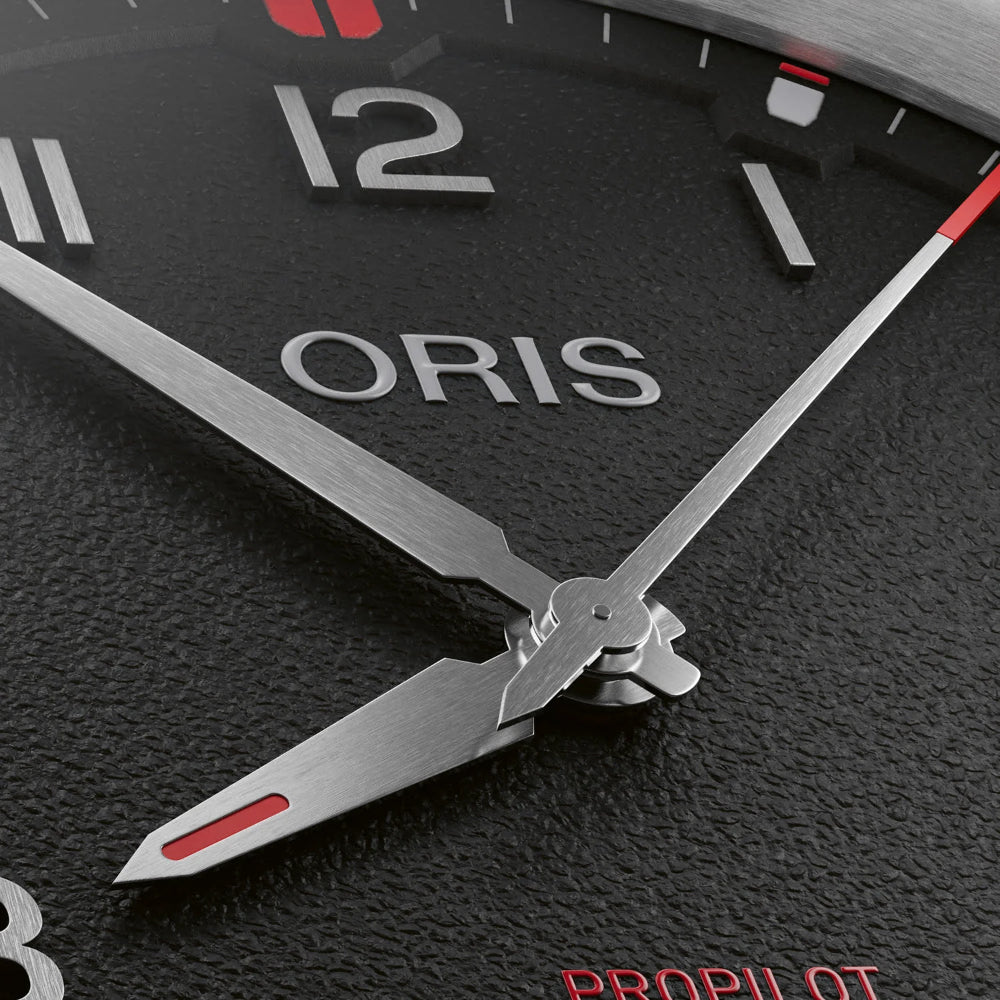 Close-up of an Oris watch dial and hands, showing a textured black face, silver metal hands, white numerals, and small red accents.