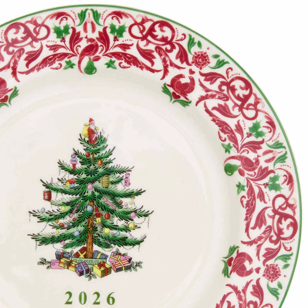 A close-up of a white ceramic plate featuring a red and green ornate floral border and a central illustration of a decorated Christmas tree with presents and the year 2026 printed below.