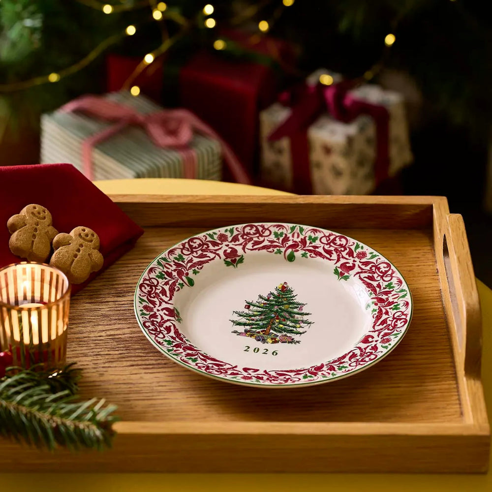 A white ceramic Christmas plate with a red holly border and green tree design, displayed on a wooden tray with holiday decorations and wrapped gifts in the background.