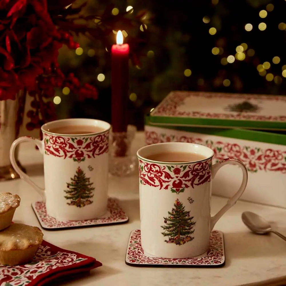 Two white ceramic mugs with red ornate borders and green Christmas tree illustrations, set on matching coasters on a table with a candle, blurred holiday lights, and a gift box in the background.