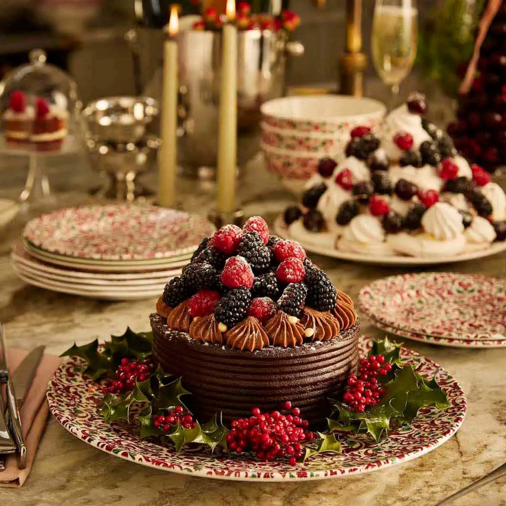 A festive holiday dessert display on a decorated table, featuring a chocolate cake topped with fresh raspberries and blackberries, surrounded by holly, alongside meringues and other seasonal sweets.