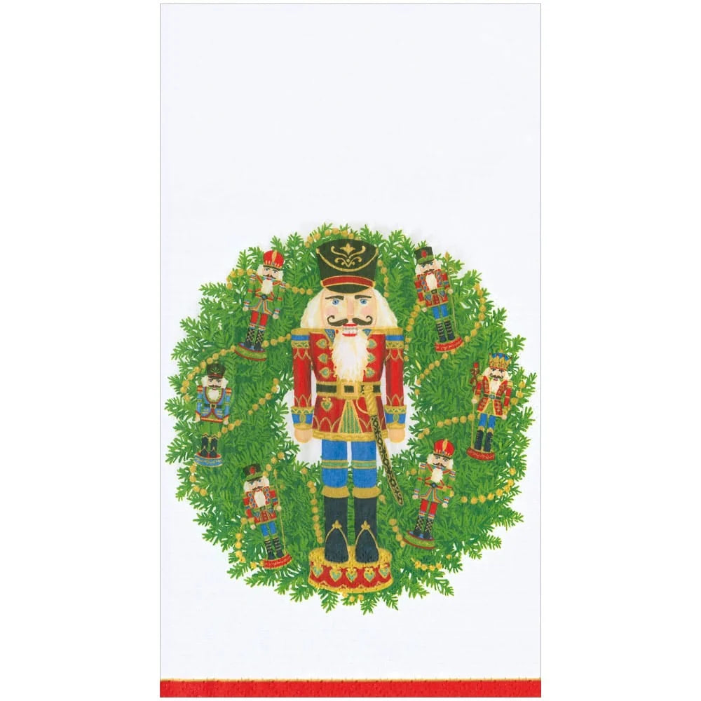 A white napkin featuring a colorful Christmas nutcracker illustration centered within a green wreath, with small nutcracker figures around it and a red stripe along the bottom edge.
