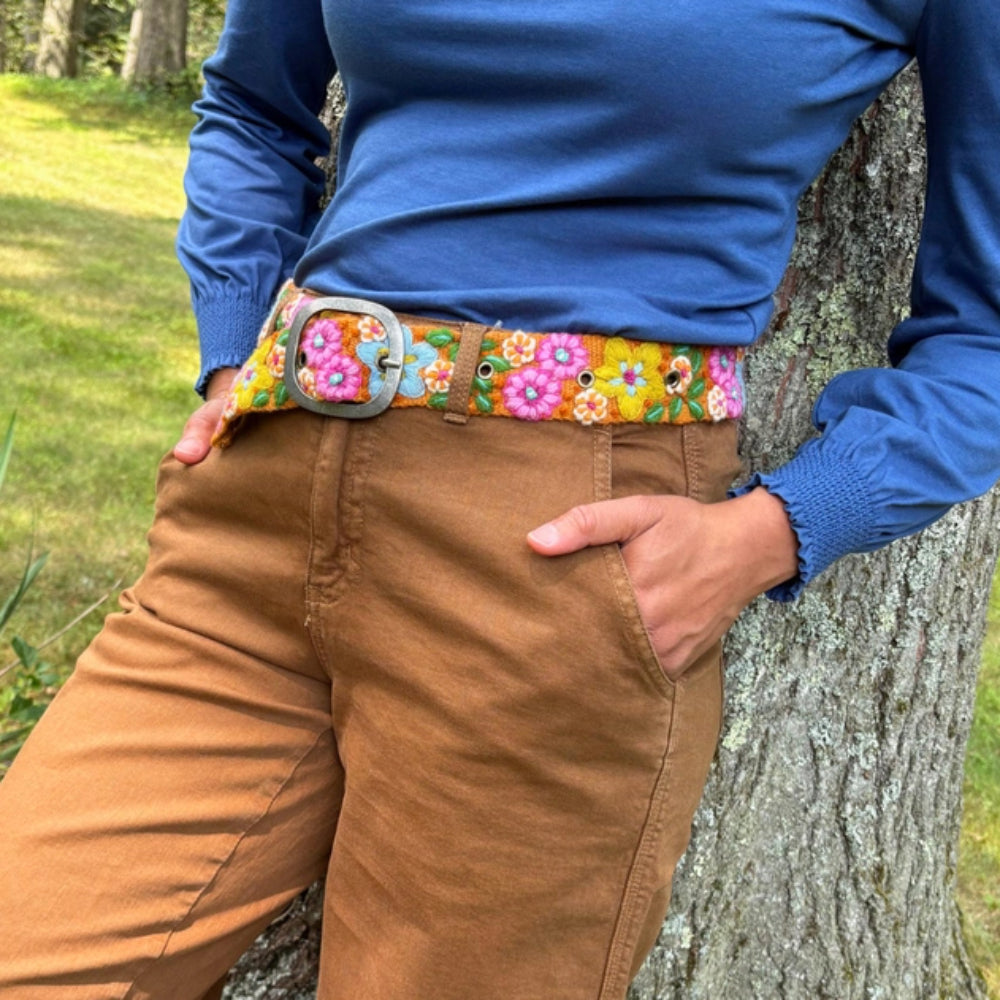 Person wearing a colorful floral belt with a blue shirt and brown pants, leaning against a tree.