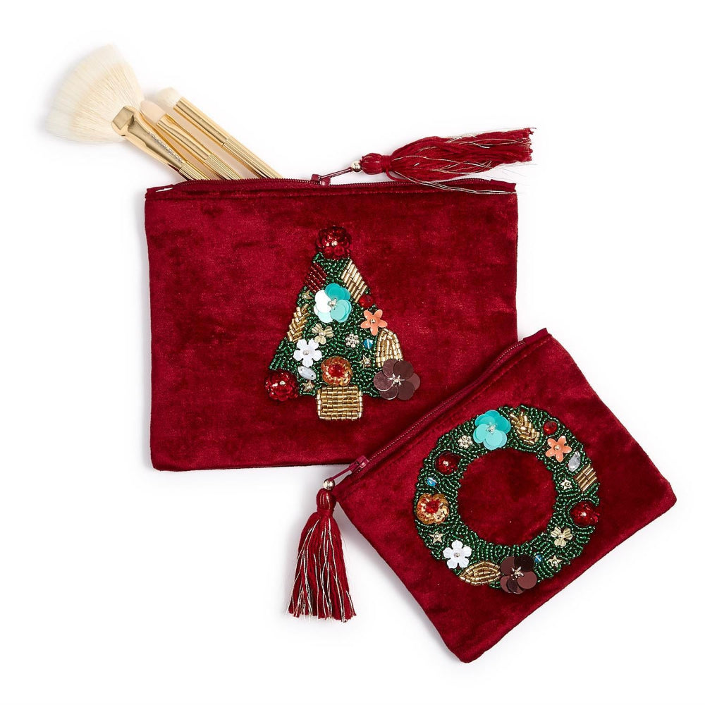 Two red velvet cosmetic pouches with embroidered holiday motifs, one featuring a Christmas tree and the other a wreath, shown on a white background with makeup brushes tucked into the top of the larger pouch.