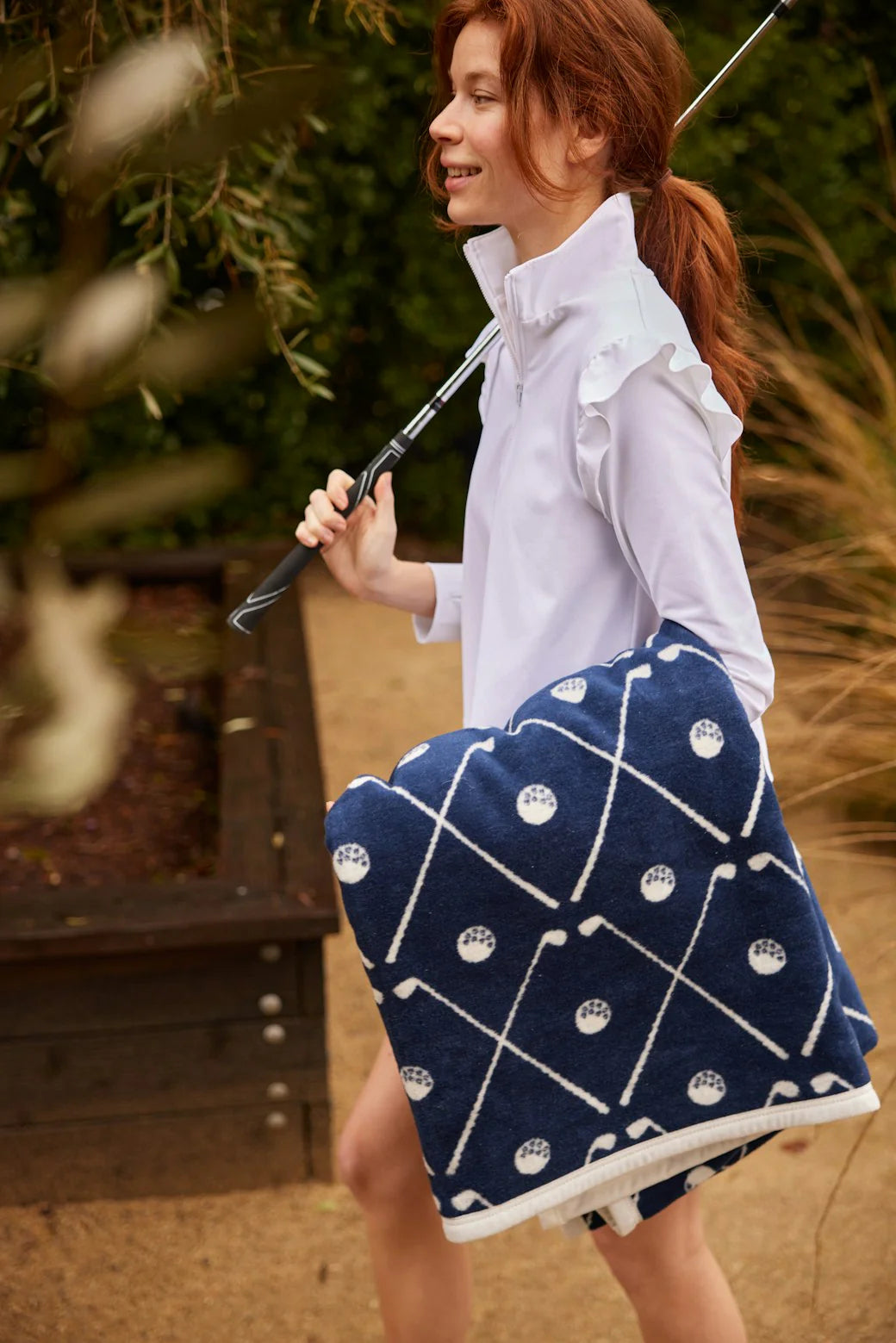 Woman holding a golf club with a blue patterned golf blanket over her shoulder outdoors.