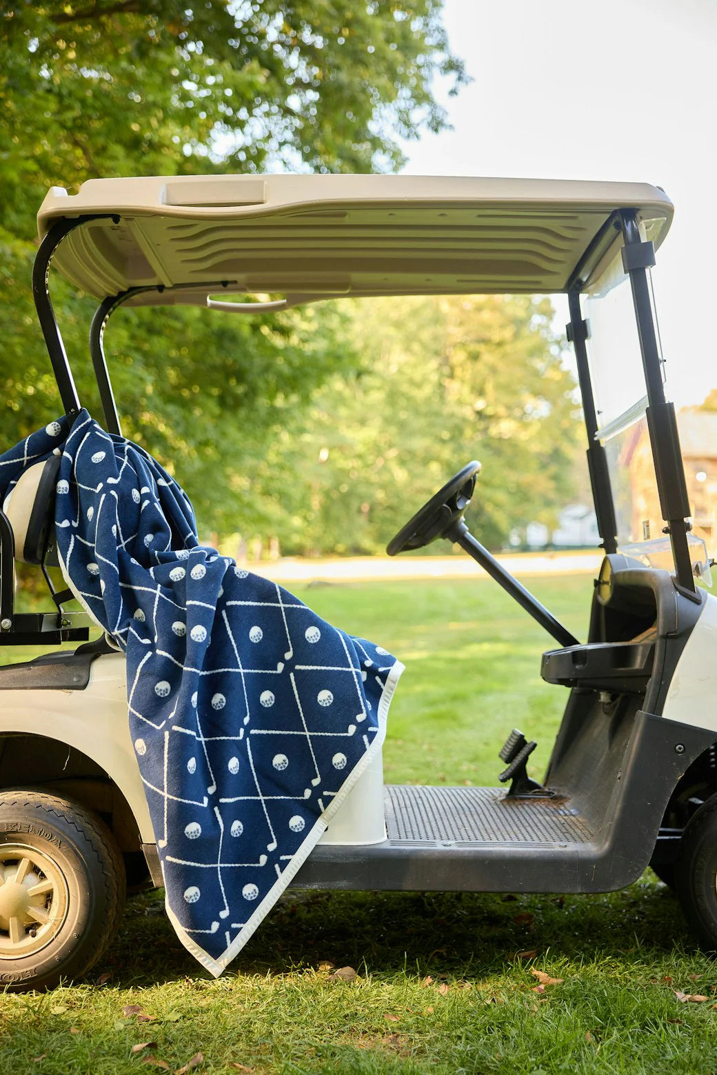 Navy blue golf blanket with white golf balls and clubs pattern draped over a golf cart on a grassy area.