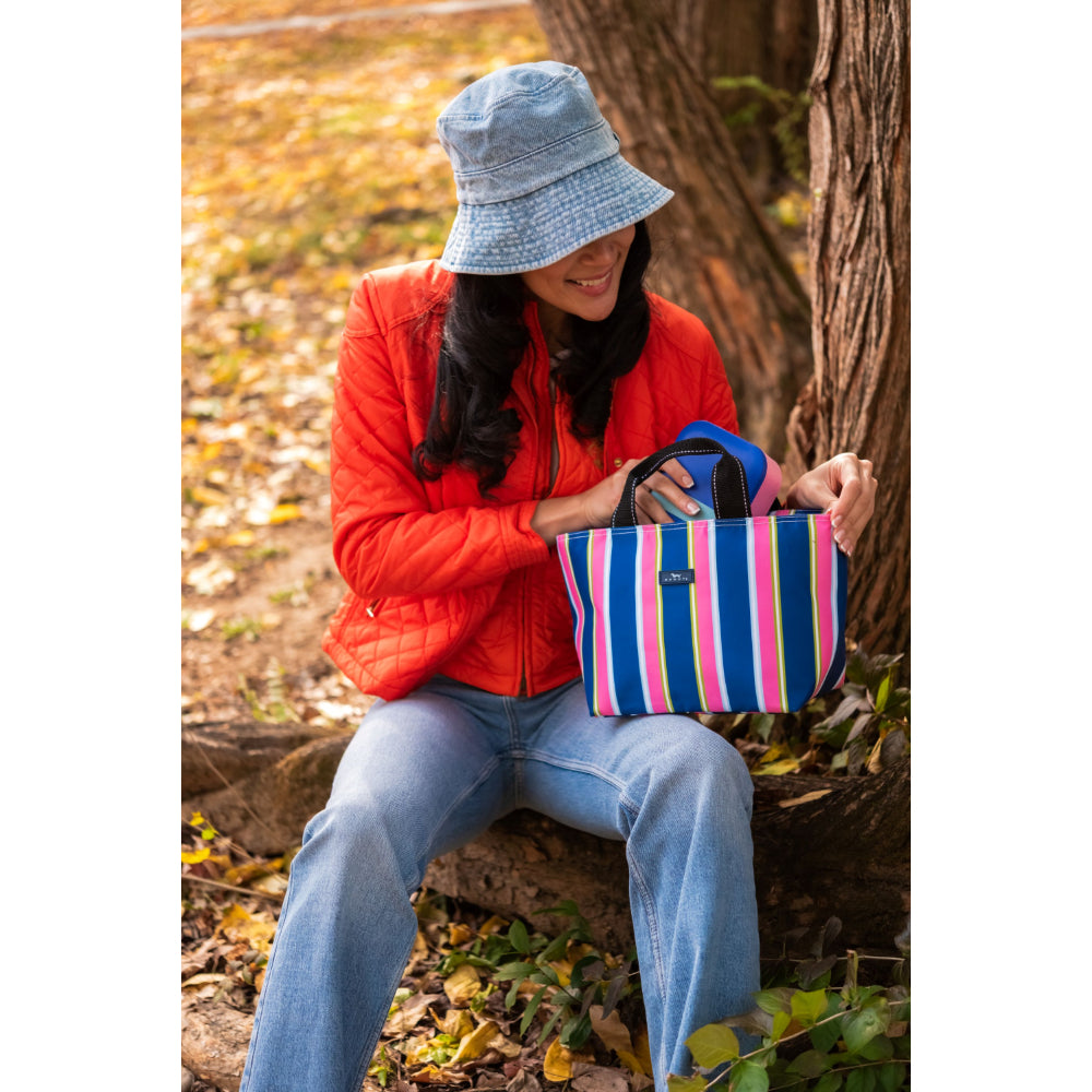 Person sitting outdoors holding a striped bag, wearing a red jacket and blue bucket hat.