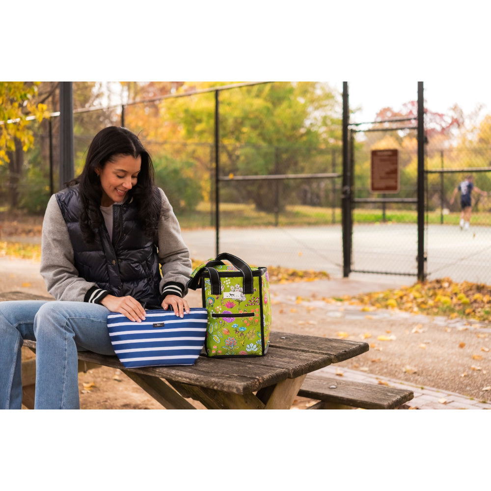 Woman sitting at a picnic table with two bags, one striped and one floral, in an outdoor setting.