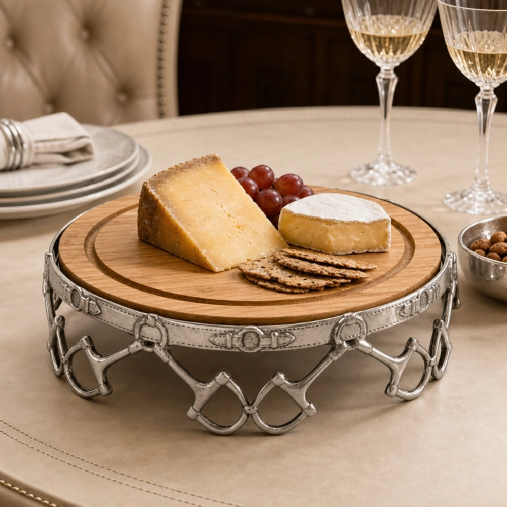 A round wooden cheese board with a decorative silver metal stand, styled on a dining table with cheese, crackers, grapes, and wine glasses in the background.