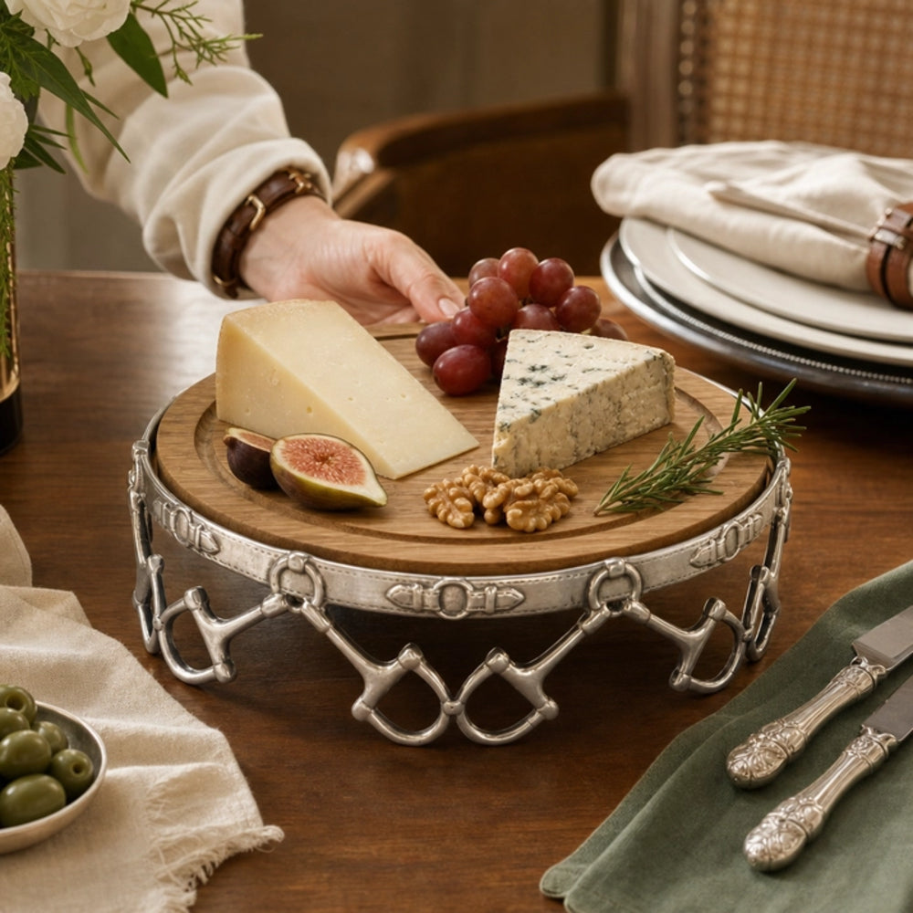 A round wooden cheese board on a silver metal stand, styled with cheese wedges, grapes, a fig, walnuts, and rosemary on a wooden dining table. A person's hand is reaching toward the cheese, and surrounding tableware including plates, cutlery, and olives are visible.