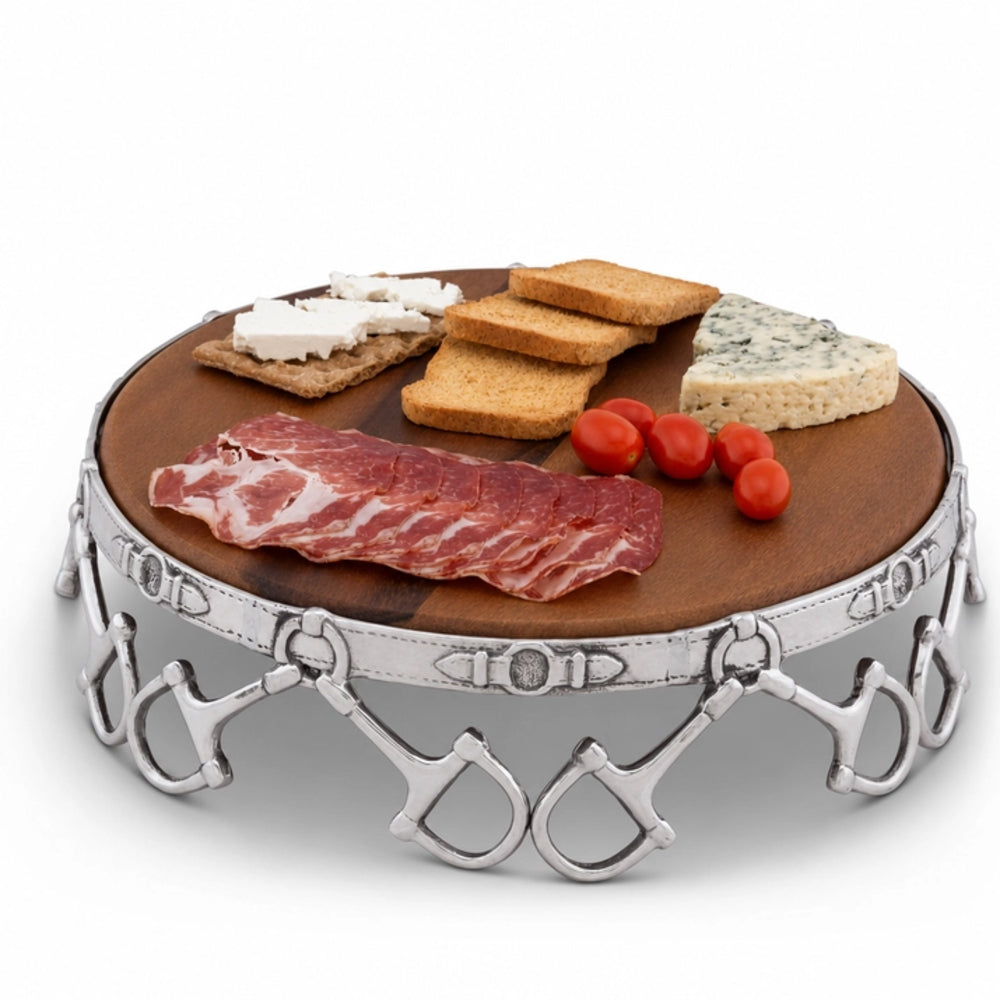 Round charcuterie board with a dark wood top and a silver metal frame decorated with horsebit details, shown filled with crackers, cheese, salami, and cherry tomatoes on a white background.