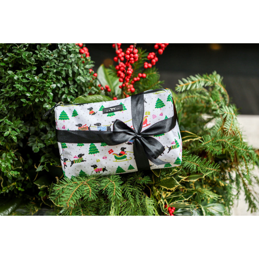 A small gift box wrapped in white paper with a Christmas dog print, tied with a black ribbon and bow, nestled among green pine branches and red berries.