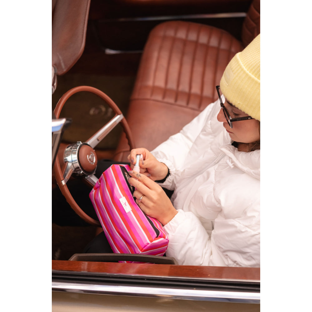 A person wearing a white puffer jacket, yellow beanie, and glasses sits inside a vintage car with brown leather seats, holding a pink and orange striped zippered pouch.