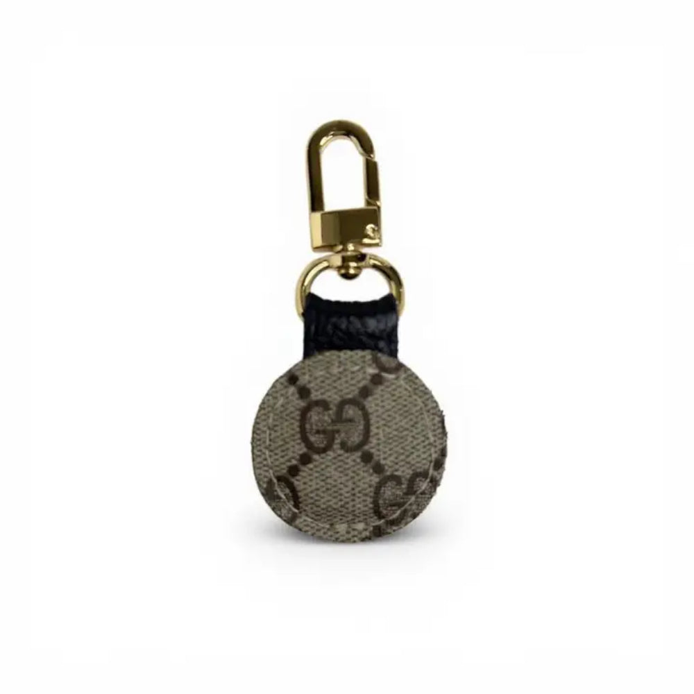 A circular Gucci keychain or bag charm made of beige and brown GG Supreme canvas with a black leather loop and a gold-tone metal clasp, shown on a plain white background.