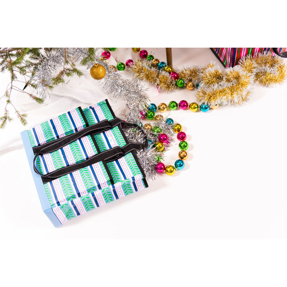 A wrapped Christmas gift box with a blue, green, and white striped pattern and black ribbon handles, styled on a white surface with festive tinsel, ornaments, and other wrapped presents.