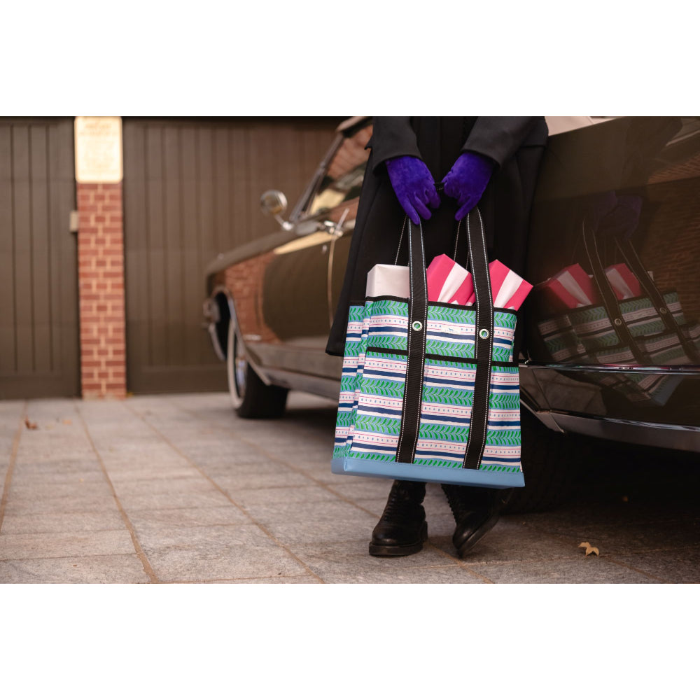 A person wearing a black coat and purple gloves holds a striped tote bag with green, white, and blue wavy patterns and black handles, standing next to a vintage car in a driveway.