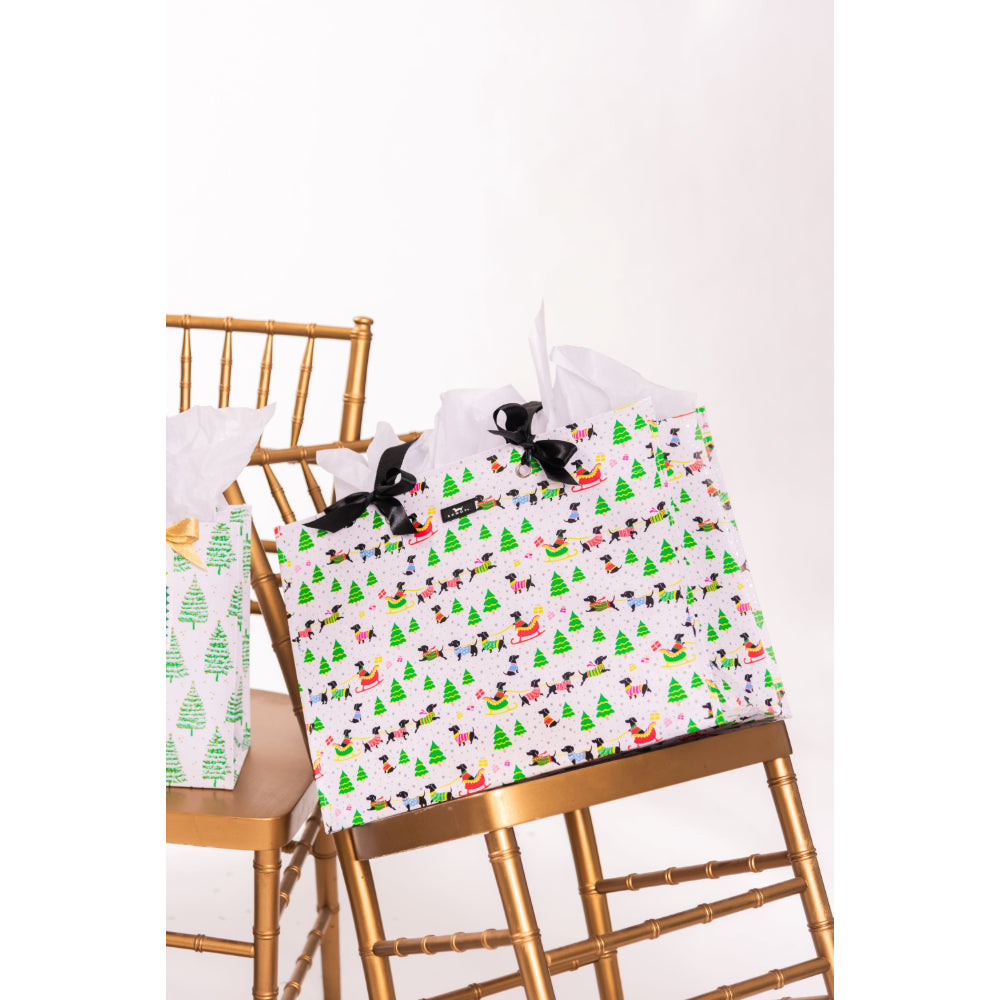 A white gift bag with a colorful Christmas-themed pattern of trees, sleighs, and snow globes, styled on a gold chair next to a matching gift box.