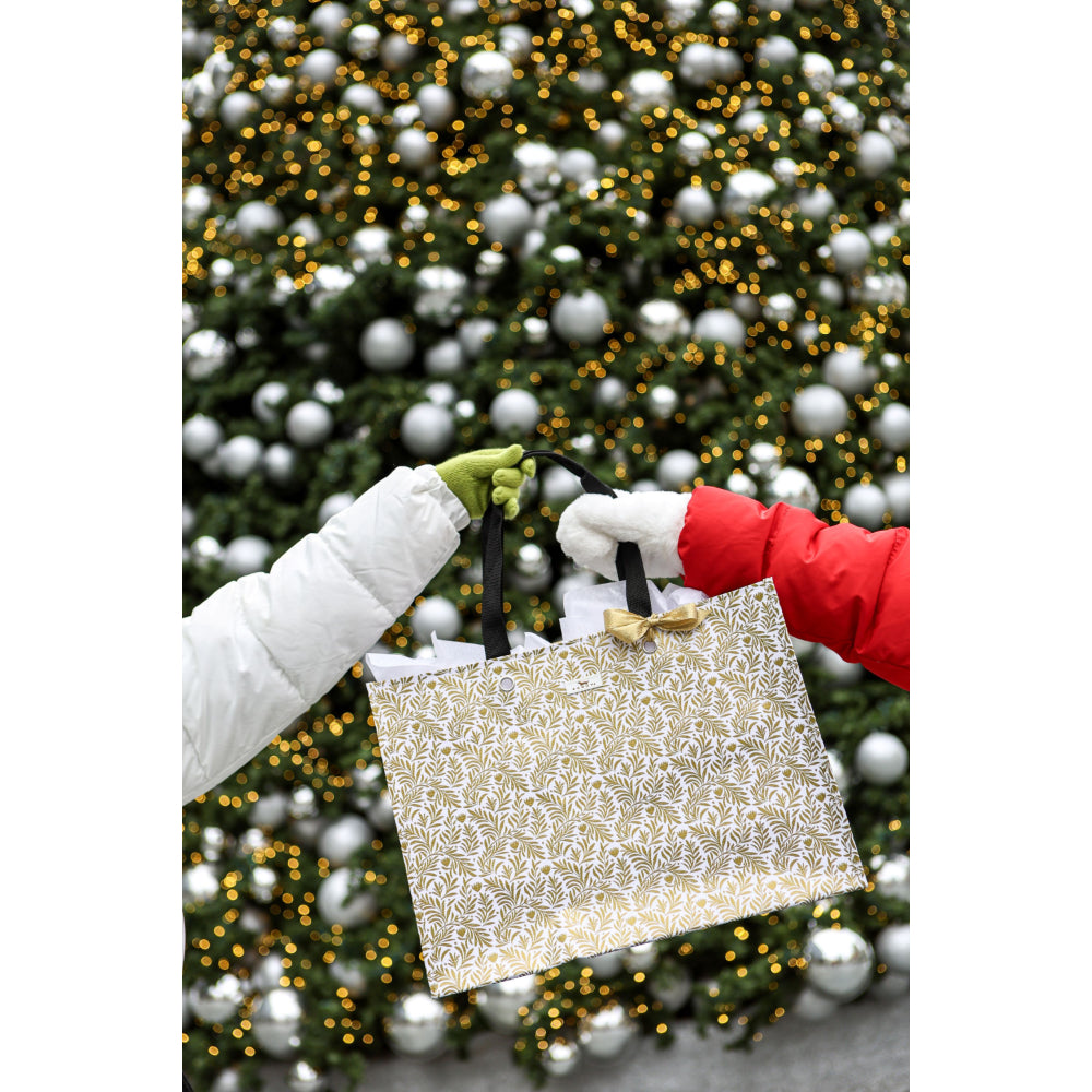 A person holds a large metallic silver gift bag with black handles in front of a decorated Christmas tree with silver ornaments and warm yellow lights. One hand wears a white puffer jacket and green glove, and the other wears a red sleeve and white glove.