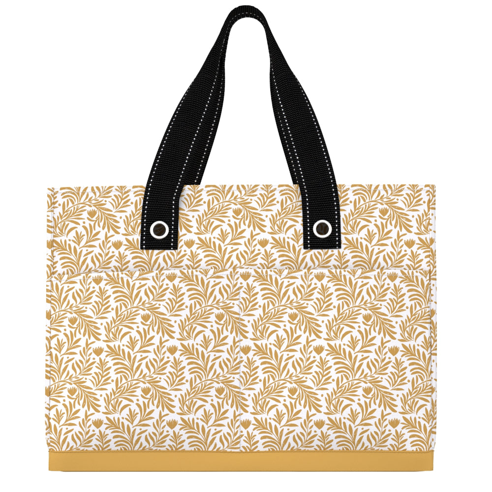 A tote bag with a white background and a repeating gold botanical leaf pattern, featuring black shoulder straps with white contrast stitching and silver grommets.