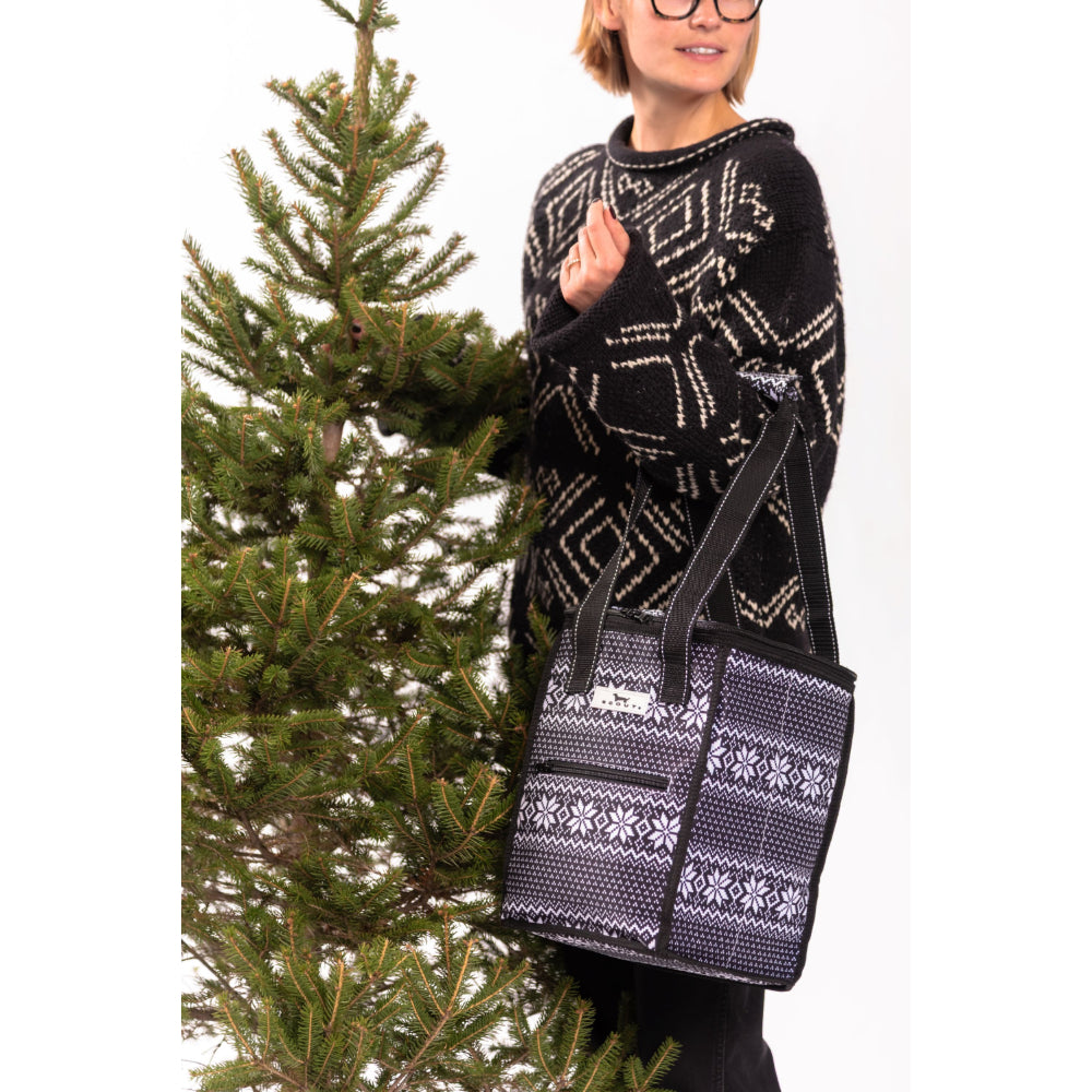 A woman wearing a black and white patterned knit sweater stands next to a Christmas tree, holding a black tote bag with a white Nordic snowflake pattern.