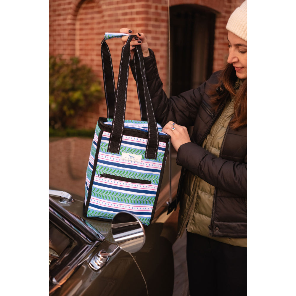 A woman in a black puffer jacket and cream beanie holds up a striped tote bag with blue, green, white, and purple geometric patterns and black handles, standing next to a black car.