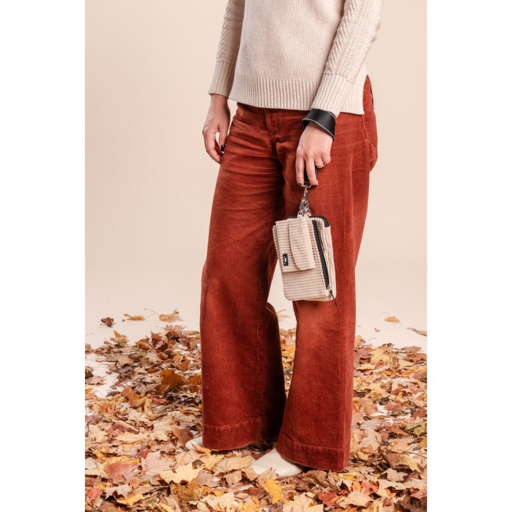 Person wearing rust-colored pants and a beige sweater, standing on a leaf-covered ground.