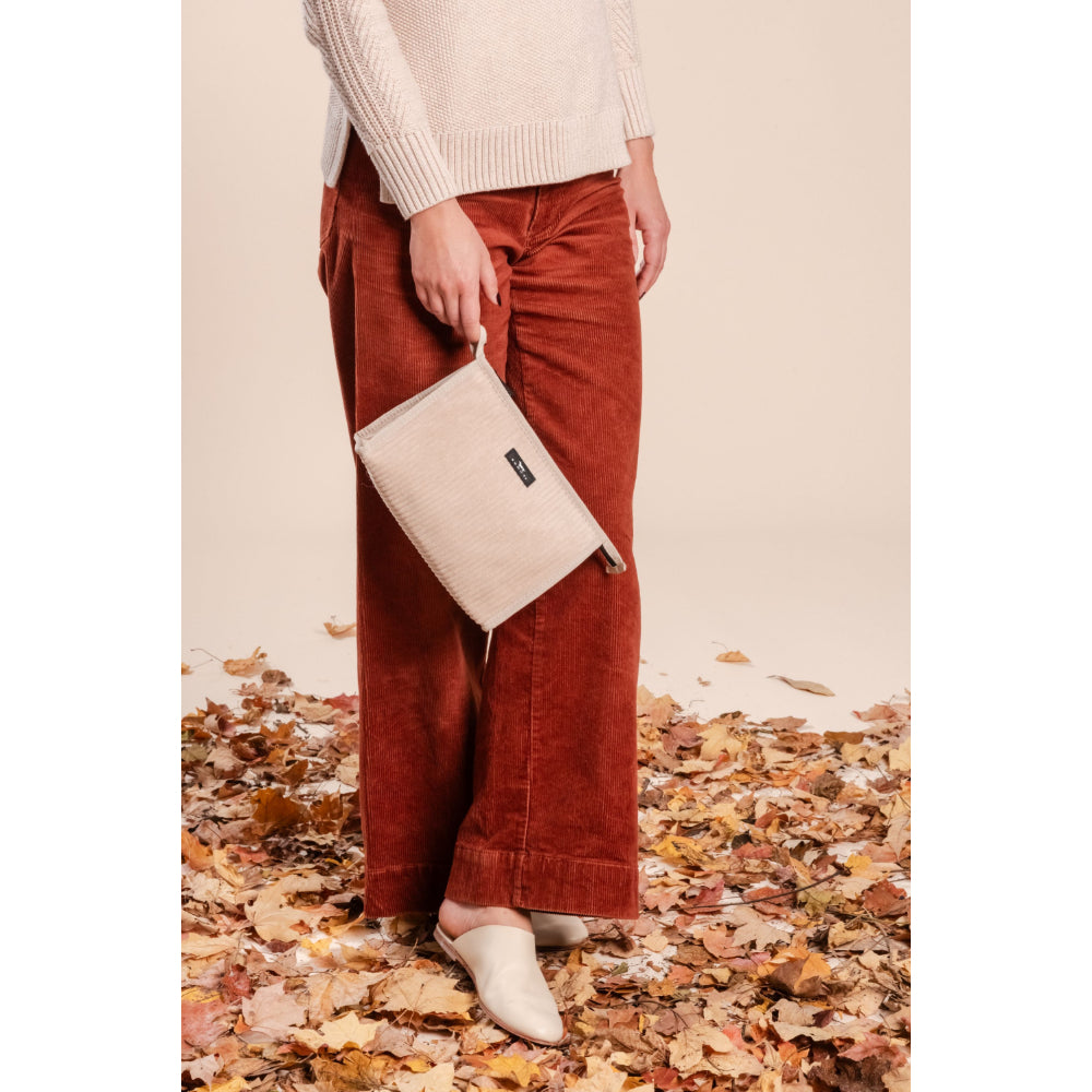 Person wearing rust-colored pants and a beige sweater, holding a beige clutch on a leaf-covered ground.