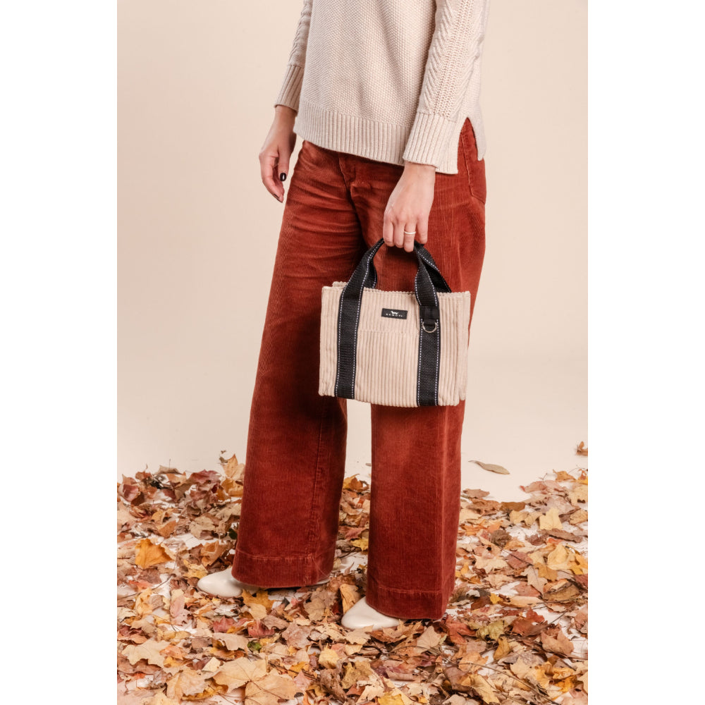 Person wearing rust-colored pants holding a TAN bag on a leaf-covered ground.