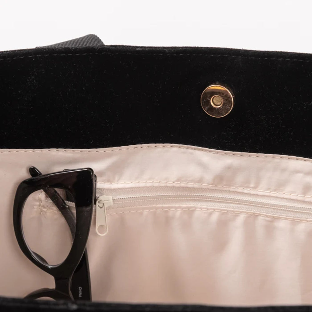 Close-up of the interior of a black bag with a cream-colored nylon lining, featuring a gold-tone snap button, a zippered pocket, and a pair of black eyeglasses resting inside.