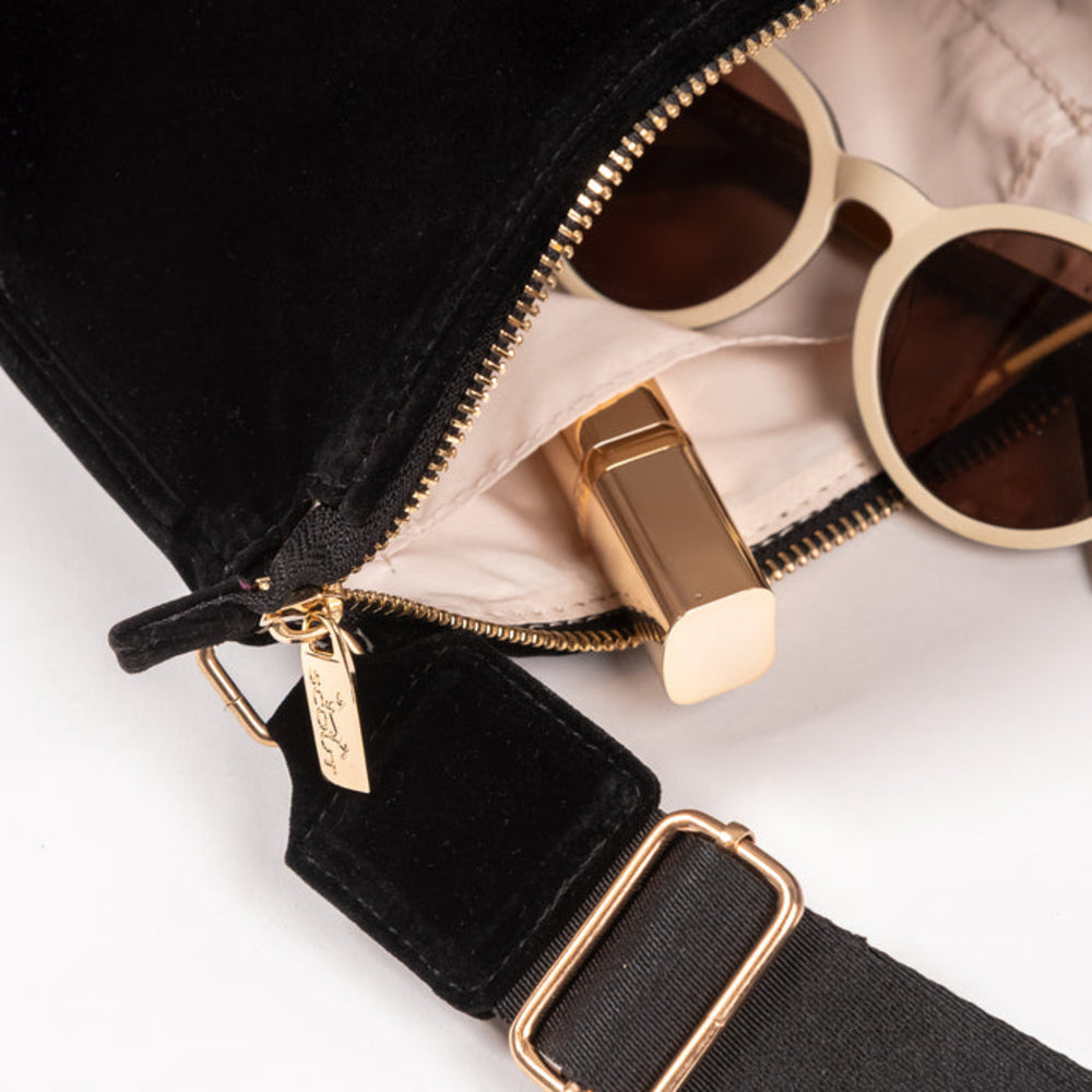 Close-up of a black velvet crossbody bag with gold-tone hardware, shown open with a cream lining and sunglasses resting inside. The zipper pull and strap buckle are visible against a clean white background.