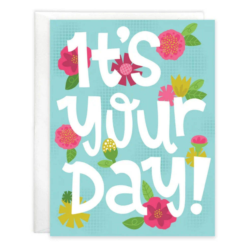 A greeting card with a white envelope behind it. The card features the phrase 'It's your Day!' in large white stylized lettering on a light blue background, decorated with pink, yellow, and green flowers and leaves.