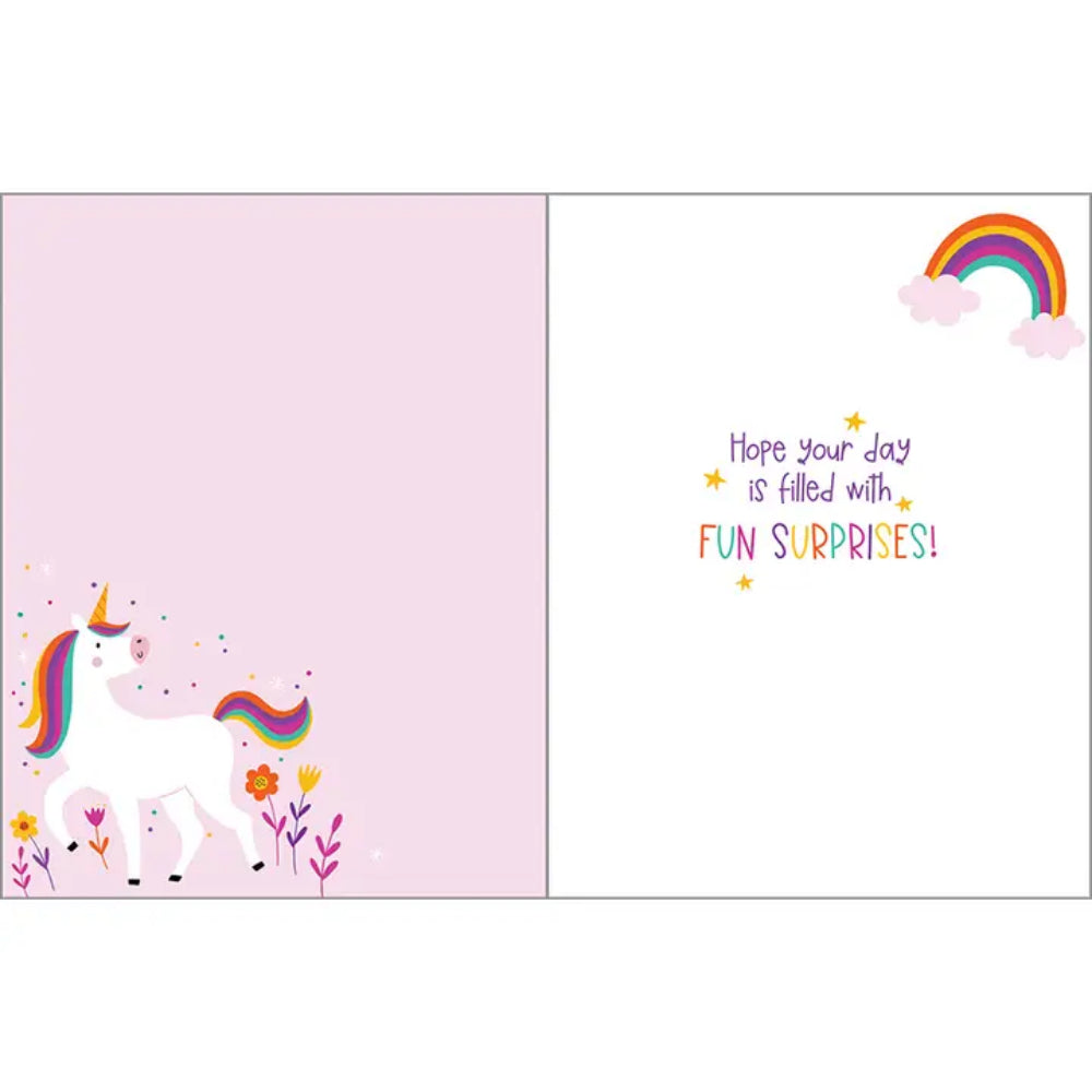 A children's surprise gift card shown open, with a pastel pink left panel featuring a white unicorn, rainbow mane, flowers, and confetti, and a white right panel with the message 'Hope your day is filled with FUN SURPRISES!' plus a small rainbow and clouds in the top right.