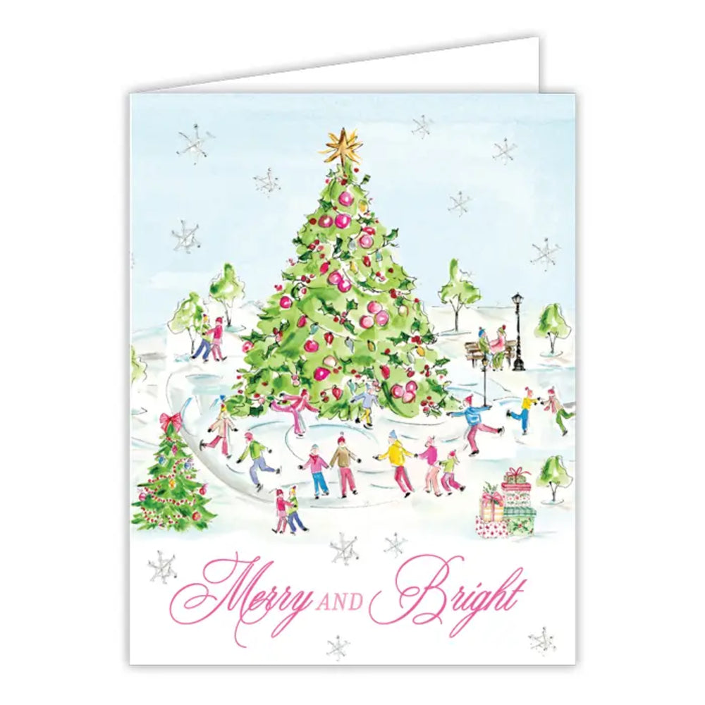 A festive greeting card featuring a watercolor-style illustration of people ice skating around a decorated Christmas tree in a snowy park. The card reads 'Merry and Bright' in pink script at the bottom, with a white envelope partially visible behind it on a white background.