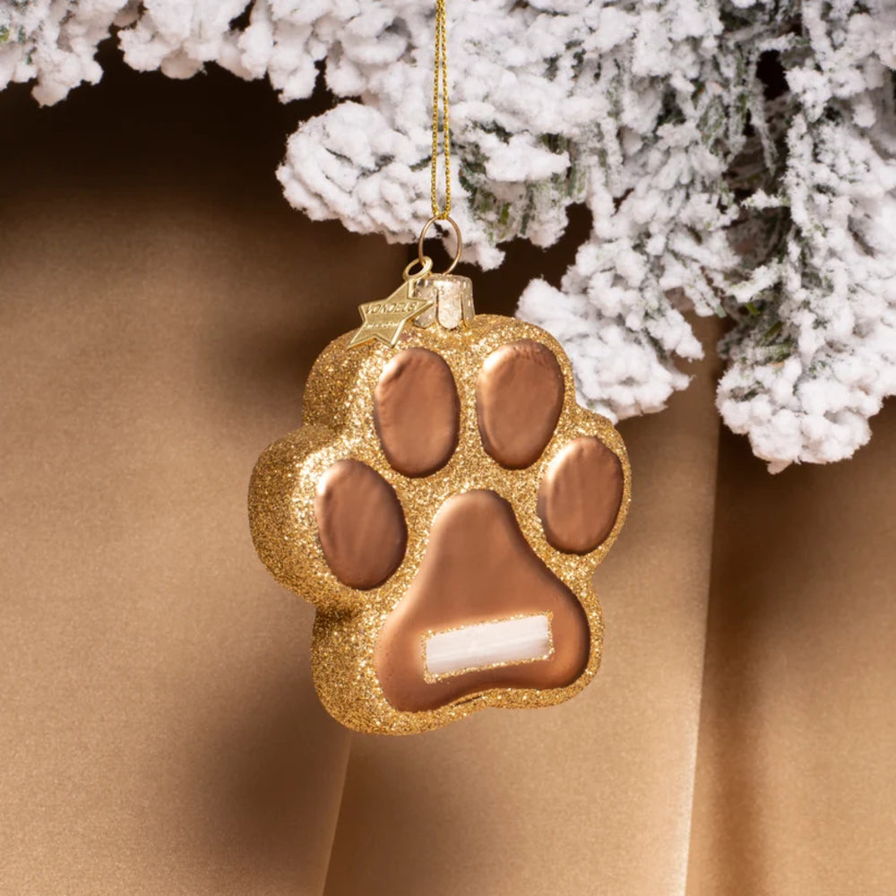 A gold glitter paw-shaped Christmas ornament with metallic rose-gold colored pads and a white accent, hanging from a gold string against a warm brown background with frosted white branches at the top.