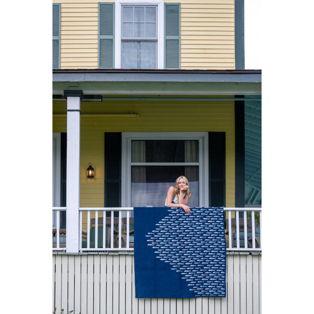 Woman hanging a blue blanket with white patterns on a balcony of a yellow house.